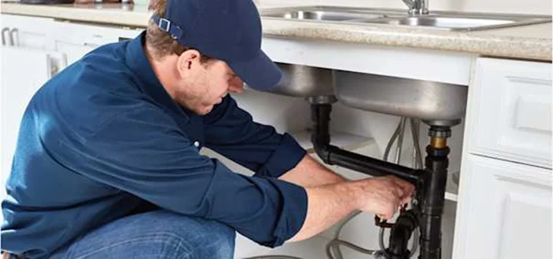 A plumber repairs a kitchen sink, kneeling beside the cabinet with tools and plumbing parts visible.